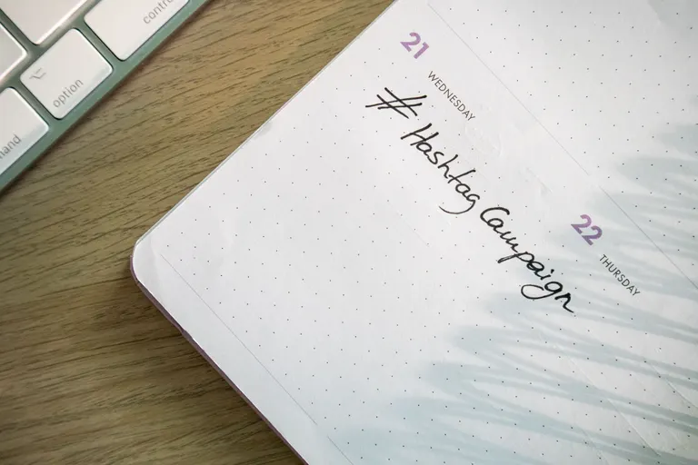 An open planner with a hashtag campaign note handwritten next to a keyboard, used for planning a marketing send